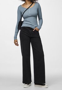 Kvinna i en ljusblå långärmad ribbad topp, svarta vida jeans, svart axelremsväska och svart-vita plattformssneakers, stående.