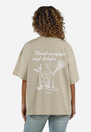 Άτομο με σγουρά μαλλιά που φοράει μπεζ μπλουζάκι με λευκή εκτύπωση κοκτέιλ και το κείμενο «Χρειάζομαι καλοκαίρι και ποτά» στην πλάτη.