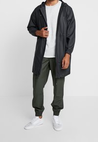 Lekka czarna wodoodporna kurtka z kapturem, zapinana na zamek, z elastycznymi mankietami. W komplecie z oliwkowozielonymi spodniami i białymi trampkami.