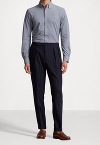 Mężczyzna w jasnoniebieskiej koszuli zapinanej na guziki, granatowych spodniach w prążki z mankietami i brązowych skórzanych mokasynach, stojący na jednolitym tle.