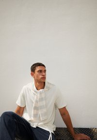 T-shirt beige clair avec un col châle, associé à une écharpe rayée crème et grise drapée autour du cou, jeans bleu foncé et une surface texturée.