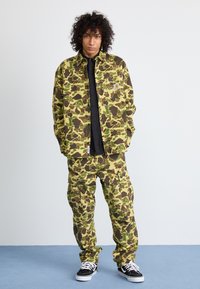 Camouflage-patterned jacket and pants in green, brown, and tan; cargo pockets; paired with black shirt and sneakers; relaxed fit.