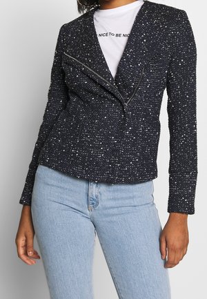 Woman wearing a speckled black textured zip-up jacket over a white shirt with "Nice to be nice" text and light blue jeans.