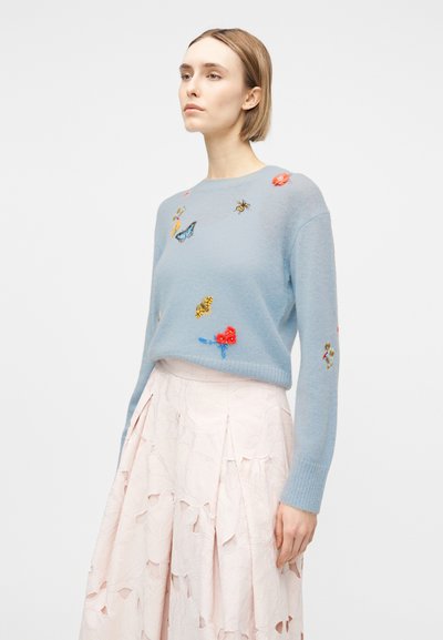 Γυναίκα με κοντά μαλλιά που φοράει ένα ανοιχτό μπλε πουλόβερ με κεντημένες πεταλούδες και λουλούδια, συνδυασμένο με μια ανοιχτό ροζ υφασμάτινη φούστα.
