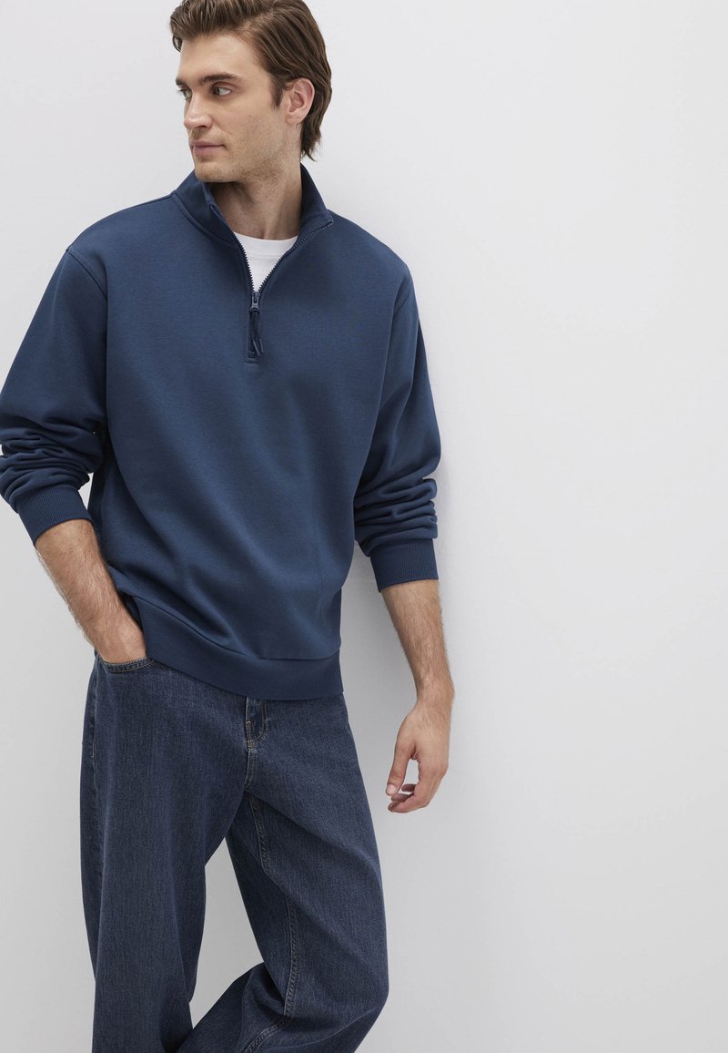 Marineblaue Zip-Hoodie mit langen Ärmeln, weichem Material und gerippten Bündchen. Kombiniert mit lockeren, dunklen Jeans.