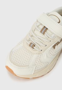 Lettvekts krem atletisk sko med mesh-overdel, beige detaljer, rund tå-design og teksturert gummisåle med en lett hevet hæl.