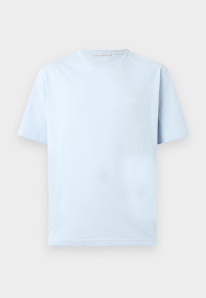 Homme aux cheveux bouclés portant une chemise bleu clair à manches courtes, un pantalon plissé gris et des chaussures noires, debout avec une main dans la poche.