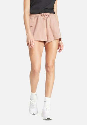 Mujer con pantalones cortos deportivos rosas con bolsillo lateral, camiseta negra, calcetines blancos y zapatillas blancas, de pie frente a un fondo blanco.