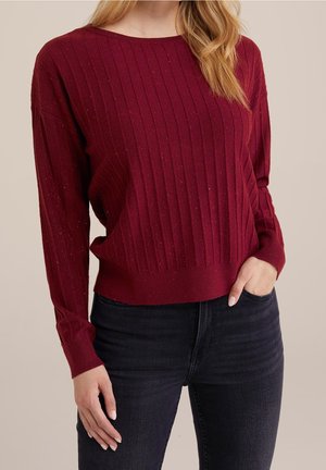 Vrouw die een bordeauxrode, geribbelde trui met lange mouwen draagt en een zwarte spijkerbroek, met één hand in haar zak en de andere naast zich rustend.