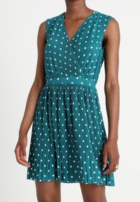 Teal pleated dress with a wrap design, sleeveless, featuring white polka dots, a fitted waistband, and a flared skirt.