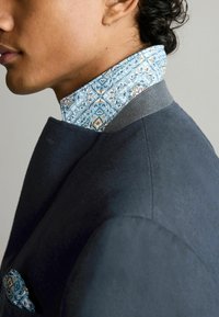Marineblaue Jacke mit strukturierter Oberfläche, ausgestattet mit einem hellblauen gemusterten Kragen und einem Einstecktuch im ähnlichen Design.
