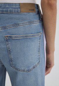 Light blue denim jeans featuring a back pocket, brown leather label with "mavi," and side detailing in pink. Close-up of back waist area.