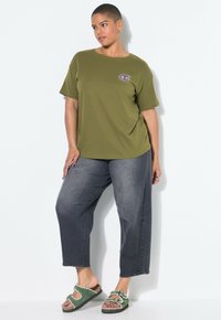 Osoba s krátkými vlasy stojí proti bílé zdi, má na sobě olivově zelené tričko s grafikou oka, vybledlé černé džíny a zelené sandály.