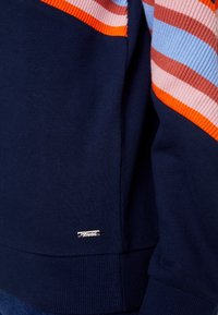 Sudadera marina con acentos a rayas multicolores en rosa, naranja y azul. Cuenta con un dobladillo acanalado y una pequeña etiqueta de logo plateado. Textura suave.