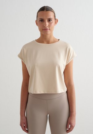 Kvinde iført en beige kropsnær crop-top og højtaljede taupe-farvede leggings, stående mod en ensartet lys baggrund.