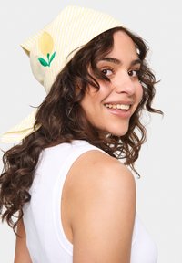 Sárga csíkos fejkendő zöld citrom és levél hímzéssel, könnyű anyagból készült, hátul megkötve, göndör hajú személy viseli.