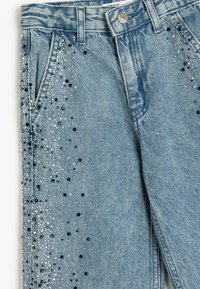 Vaaleansiniset farkut, suoralinjaiset, koristeltu metallinhohtoisilla ja mustilla strasseilla ulkopuolella koristeellisina yksityiskohtina.