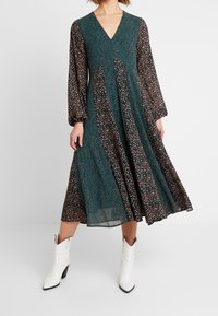 Grünes Blumendruckkleid mit langen, puffigen Ärmeln und V-Ausschnitt; knielang, aus leichtem Stoff; kombiniert mit weißen spitzen Stiefeln.