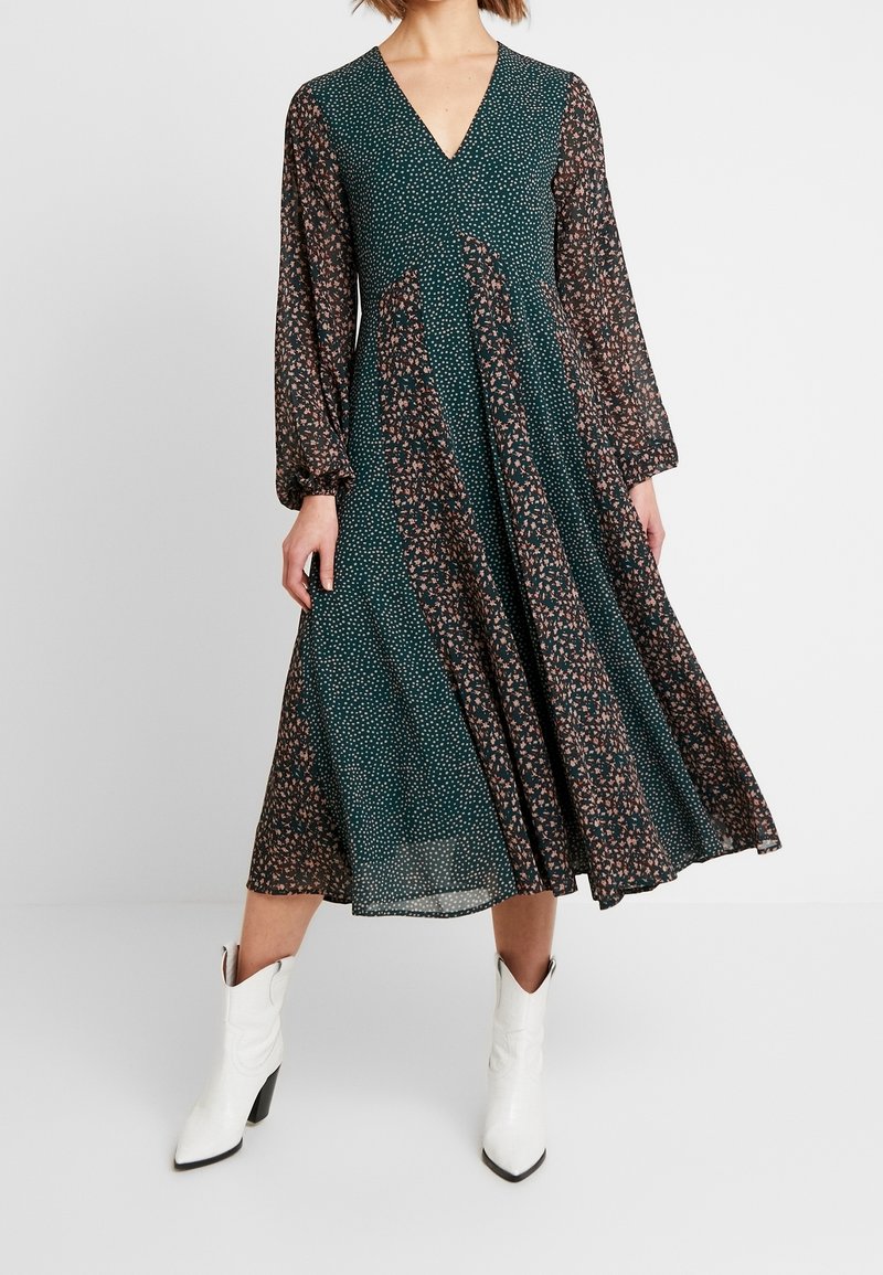 Grünes Blumendruckkleid mit langen, puffigen Ärmeln und V-Ausschnitt; knielang, aus leichtem Stoff; kombiniert mit weißen spitzen Stiefeln.