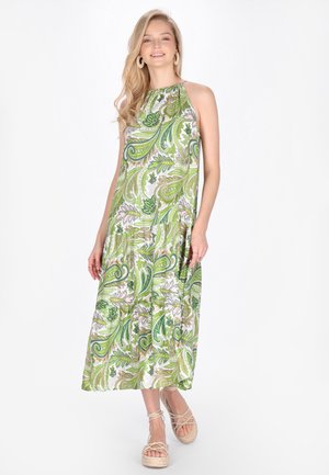 Mladá žena v zelených a bílých maxi šatech s paisley vzorem bez rukávů a béžových platformových sandálech, usmívající se a stojící na bílém pozadí.