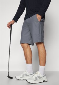 Grå idrettsshorts med teksturert overflate, sidelommer og løs passform, kombinert med hvite joggesko og en golfkølle.
