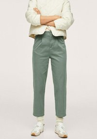 Osoba w jasnozielonych spodniach z wysokim stanem, białych trampkach z beżowymi akcentami, kremowych skarpetkach i białej pikowanej kurtce, z założonymi na siebie rękami.