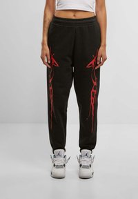 Svarte joggebukser med røde flamme-lignende grafiske design, elastisk midje, smalere ben og mykt bomullsmateriale. Brukt med hvite joggesko.