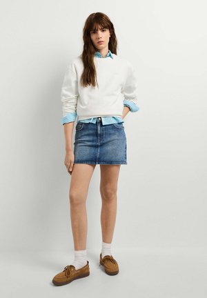 Frau in weißem Pullover über blauem Hemd, Jeansrock, weißen Socken und braunen Bootsschuhen steht mit einer Hand in der Tasche vor schlichten Hintergrund.