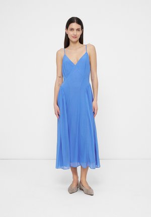 Mujer con cabello largo y oscuro, vestida con un vestido midi azul sin mangas con tirantes finos y zapatos beige planos, posando sobre un fondo blanco liso.