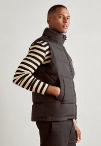 Black puffer vest with high collar and zippered front, featuring side pockets, layered over a black and beige striped long-sleeve shirt.