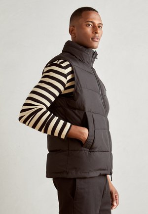 Black puffer vest with high collar and zippered front, featuring side pockets, layered over a black and beige striped long-sleeve shirt.