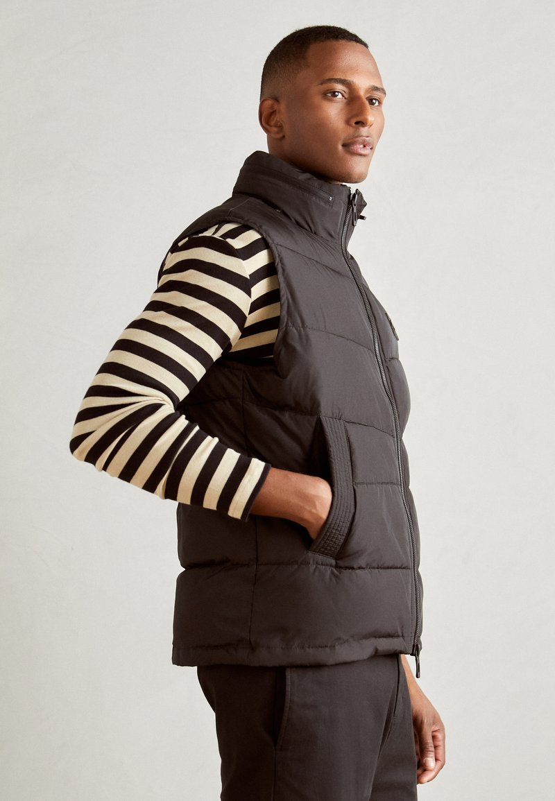 Black puffer vest with high collar and zippered front, featuring side pockets, layered over a black and beige striped long-sleeve shirt.