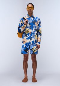 Blå sæt bestående af skjorte og shorts med blomsterprint i gule og orange nuancer. Glat stof, knap-op krave og store firkantede solbriller.