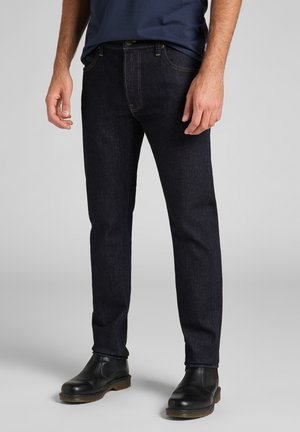 Man draagt donkerblauwe slim-fit jeans, zwarte leren enkellaarsjes en een marineblauw overhemd, staand tegen een effen grijze achtergrond.