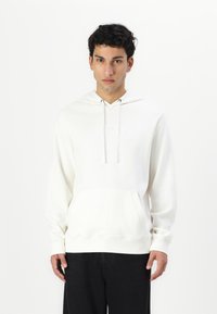 Hanorac alb dintr-un material moale, cu un buzunar frontal, șnur ajustabil și un logo minimalist pe piept; purtat cu pantaloni negri.
