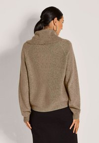 Vrouw met donker haar in een lage paardenstaart, draagt grote hoepeloorbellen, een bruin geribd coltrui en een zwarte rok, staat iets zijwaarts gedraaid.