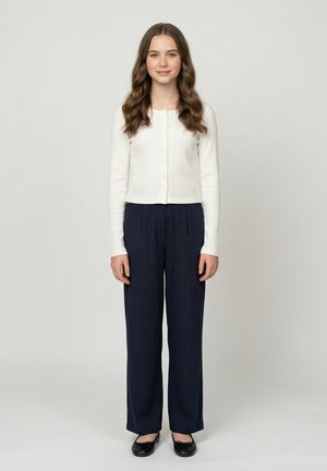 Cardigan blanc côtelé avec boutons sur le devant, associé à un pantalon large bleu marine. Chaussures plates noires complètent la tenue. Fond neutre.
