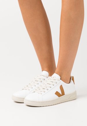 Valkoiset matalat tennarit, joissa on ruskea mokkainen "V"-logo, jalassa vaaleaa taustaa vasten.