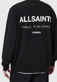 Persona con sudadera negra que lleva el texto blanco "ALLSAINTS", caracteres japoneses y "LONDON" en la parte de atrás.