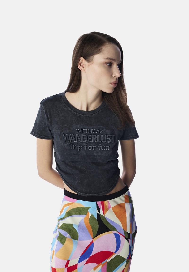 T-shirt in cotone nero con testo in rilievo "WANDERLUST Viaggio per divertirsi" e scollatura rotonda, abbinato a una gonna a fantasia astratta colorata.