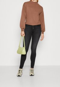 Brauner Strickpullover mit Ballonärmeln, schwarze Skinny-Jeans und eine kleine, strukturiert grüne Handtasche. Getragen mit beigen Sneakern mit schwarzen Akzenten.