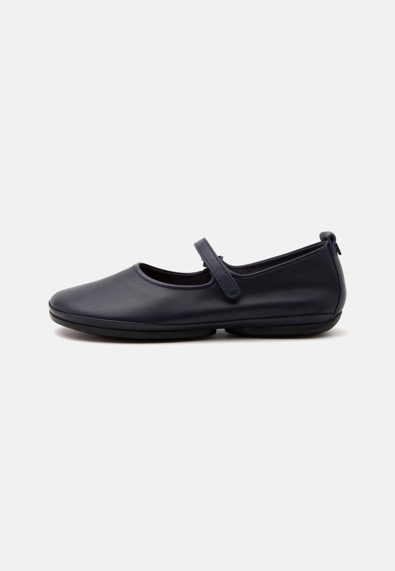 Chaussure plate Mary Jane en cuir noir avec bout arrondi, bride réglable et semelle basse en caoutchouc sur fond blanc.