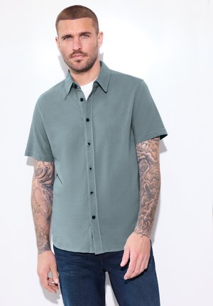 Uomo con capelli corti e tatuaggi indossa una camicia grigia a maniche corte sopra una maglietta bianca, in piedi davanti a uno sfondo bianco.