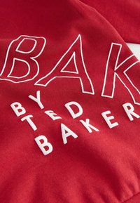 Red fabric featuring white embroidered text reading "BAKED BY TED BAKER.” The texture is smooth with a slight sheen.