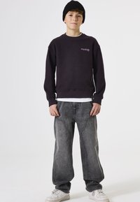 Sudadera negra con puños de canalé, que presenta "TOTAL SOLEDAD" en rosa en el pecho. Combinada con jeans grises holgados y zapatillas blancas.