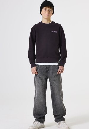 Sudadera negra con puños de canalé, que presenta "TOTAL SOLEDAD" en rosa en el pecho. Combinada con jeans grises holgados y zapatillas blancas.