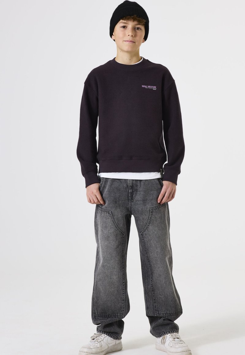 Sudadera negra con puños de canalé, que presenta "TOTAL SOLEDAD" en rosa en el pecho. Combinada con jeans grises holgados y zapatillas blancas.