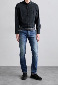 Svart skjorta med knappar, blå jeans med urtvättade partier och svarta sneakers. Outfitten har ett silverbälte spänne och en ficka på skjortan.