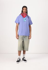 Camiseta corta de color púrpura con diseño gráfico, pantalones cortos de mezclilla verde claro, bandana roja, zapatillas negras y calcetines blancos. Textura suave.