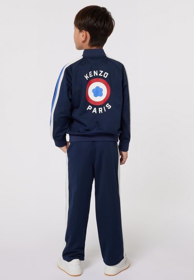 Survêtement bleu marine avec des rayures blanches et bleu clair, présentant un logo rond en rouge et bleu au dos avec le texte "KENZO PARIS" au-dessus.